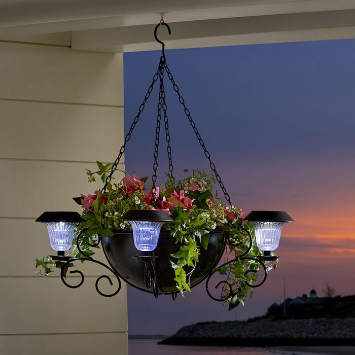 A hanging planter displays colorful flowers, illuminated by attached lights, against a scenic twilight backdrop featuring water and distant land.
