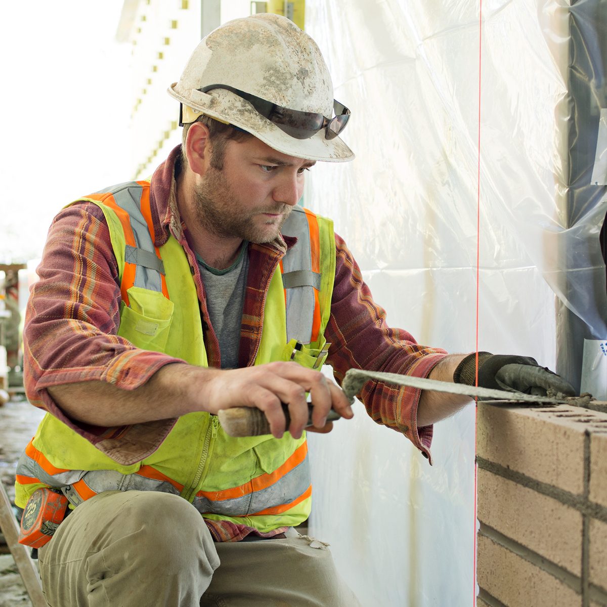 A construction worker uses a trowel to smooth masonry blocks while crouched, surrounded by plastic sheeting and tools in a partially finished building environment.