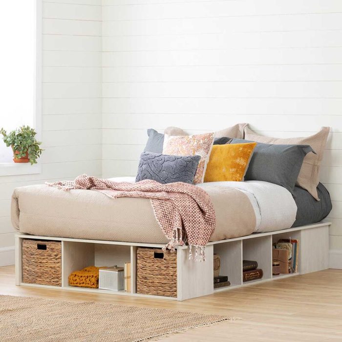 A bed with decorative pillows rests on a low platform, featuring storage baskets underneath, set against a light-colored wall and wooden floor.