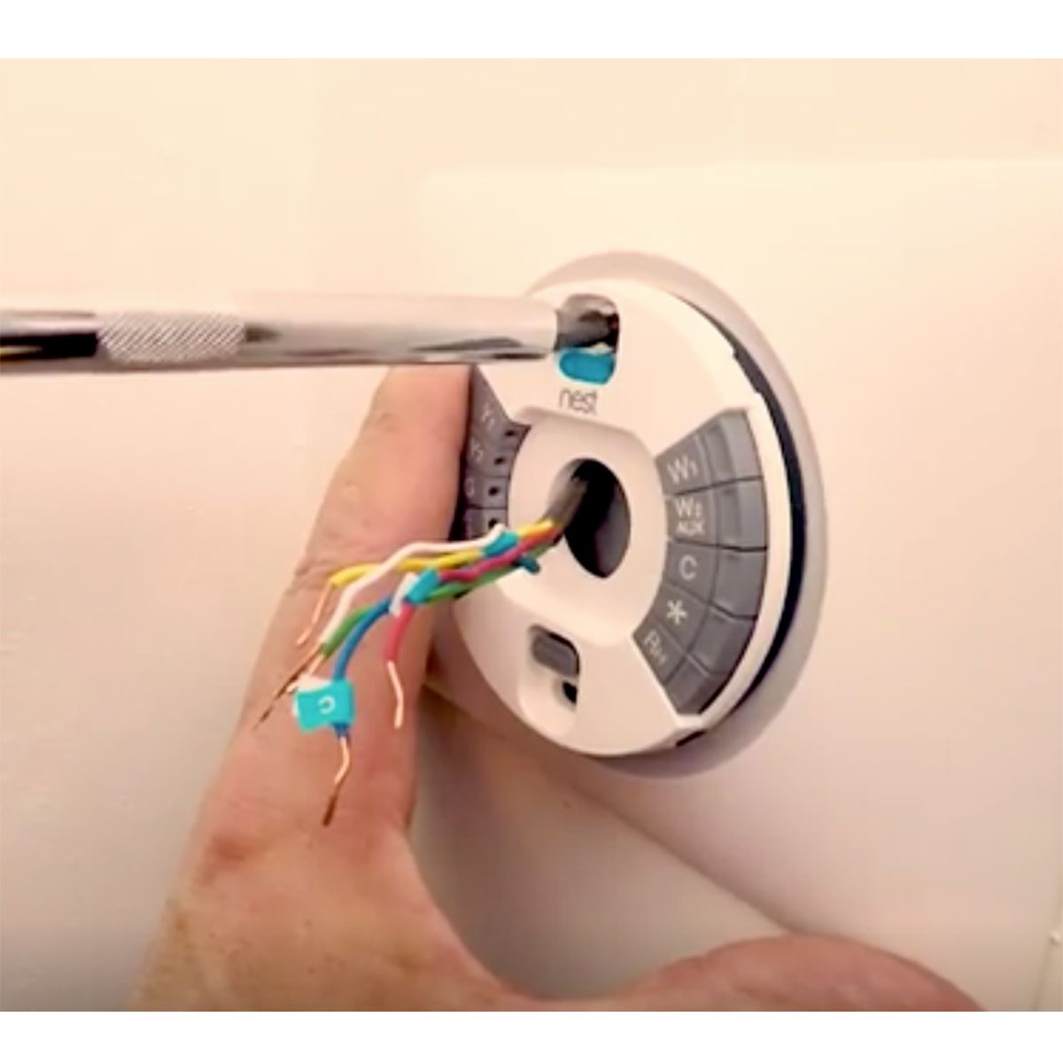 A hand holds a thermostat attachment while a screwdriver adjusts wiring connections, set against a neutral wall background.