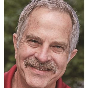 A smiling man with gray hair and a mustache is outdoors, surrounded by greenery, conveying a warm and friendly demeanor.