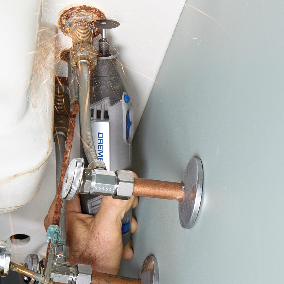 A hand holds a Dremel tool, cutting through a rusty pipe under a sink, creating sparks in a confined, utilitarian space surrounded by plumbing fixtures.