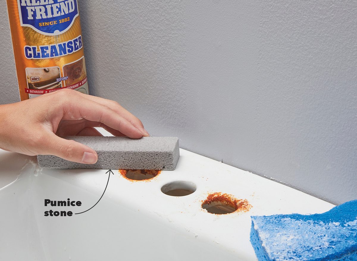 A hand holds a pumice stone, scrubbing away rust stains from a white sink, with a blue sponge nearby and cleanser visible in the background.
