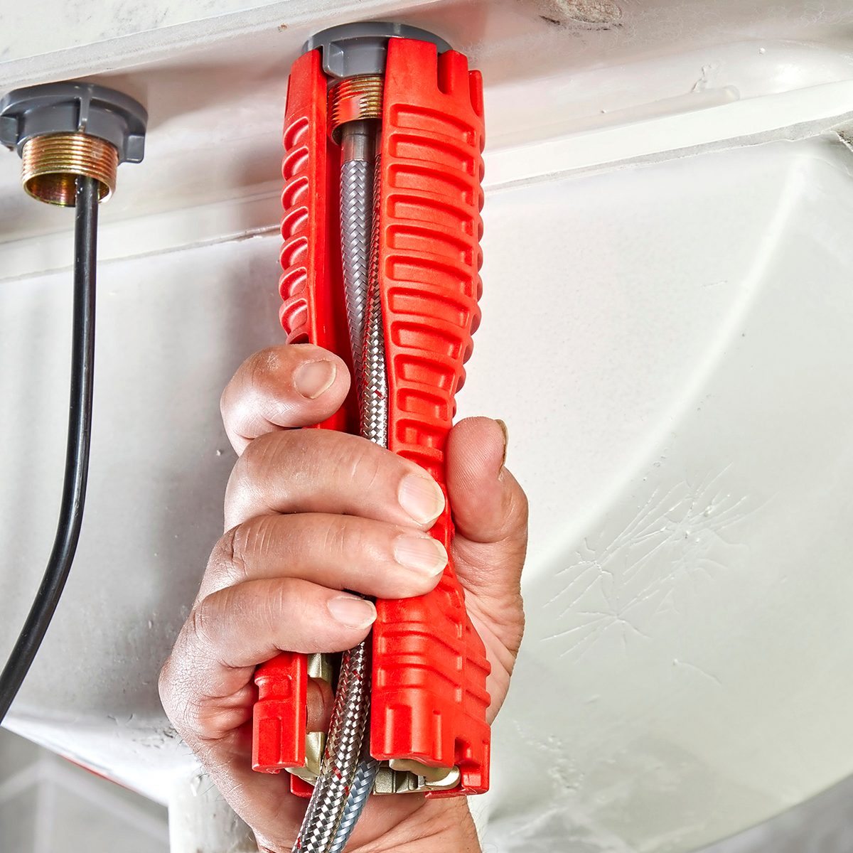 A hand holds a red tool gripping a metal hose, positioned beneath a countertop area with plumbing connections visible above.