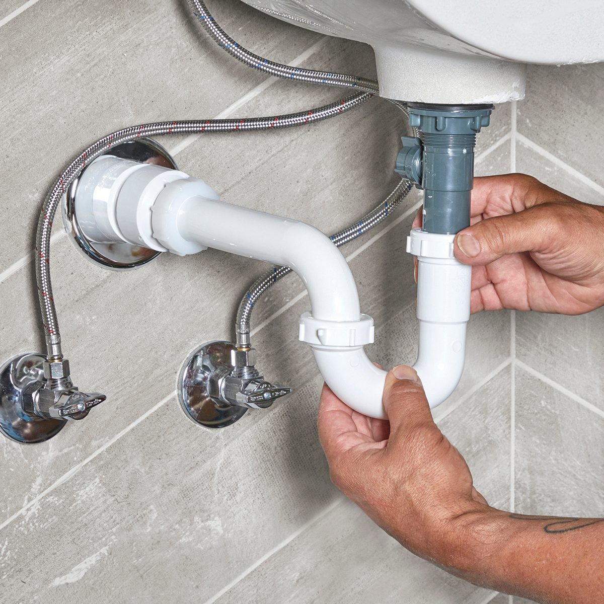 A person connects white plumbing pipes under a sink, surrounded by metal fittings and a tiled wall, indicating ongoing plumbing installation or repair.