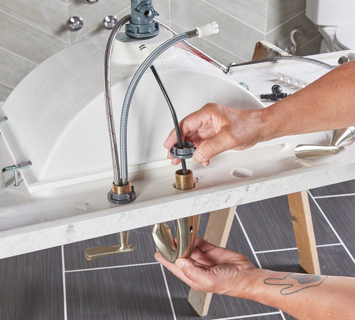 A person connects pipes beneath a sink, using tools in a bathroom with tiled walls and a basin on a stand.