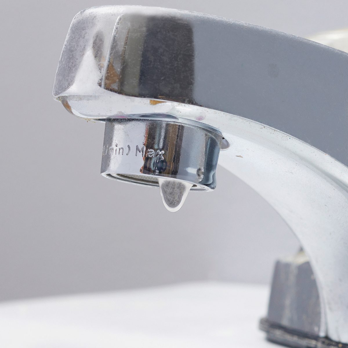 A faucet drips water from its spout, reflecting light, against a gray background, set on a white countertop.
