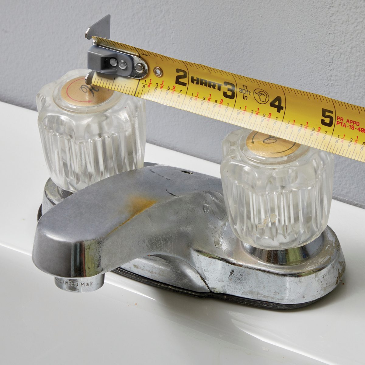 A metal faucet with clear knobs is being measured with a tape measure, positioned above a sink against a gray wall.