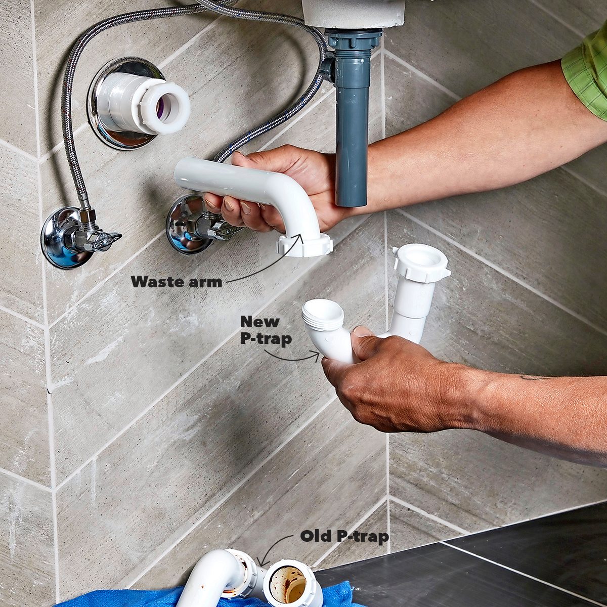 A person is replacing plumbing under a sink, holding a new P-trap and waste arm, with an old P-trap on a towel nearby.
