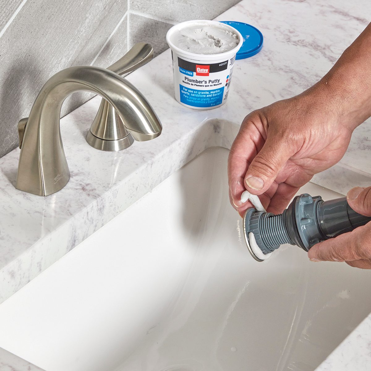 A person applies plumber's putty to a faucet attachment above a white sink, with a container of plumber's putty nearby on a marble countertop.