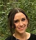 A person stands smiling among green foliage, with a natural backdrop enhancing the cheerful expression and casual attire.