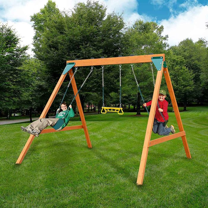 Two children swing joyfully on a wooden swing set in a grassy park surrounded by trees under a bright blue sky.