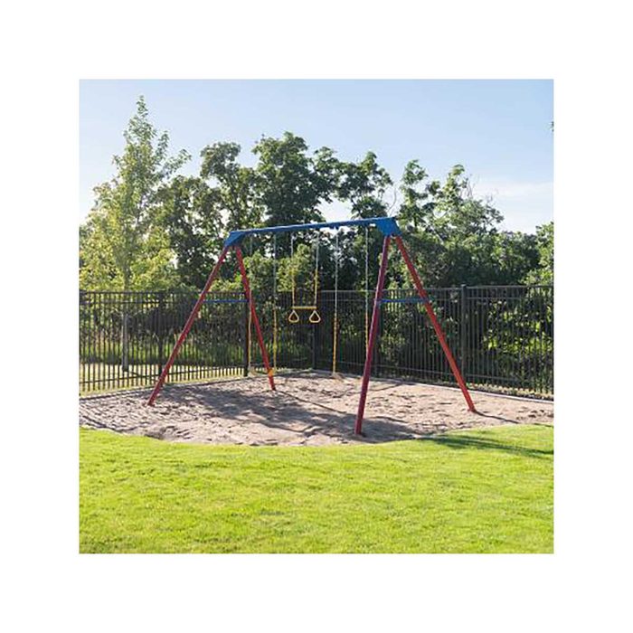 A set of swings hangs from a colorful frame, resting on sandy ground enclosed by a fence, surrounded by trees under a clear sky.