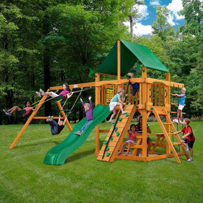 A wooden playground structure features a slide, swings, and climbing wall. Several children play energetically, surrounded by grass and trees under a clear sky.