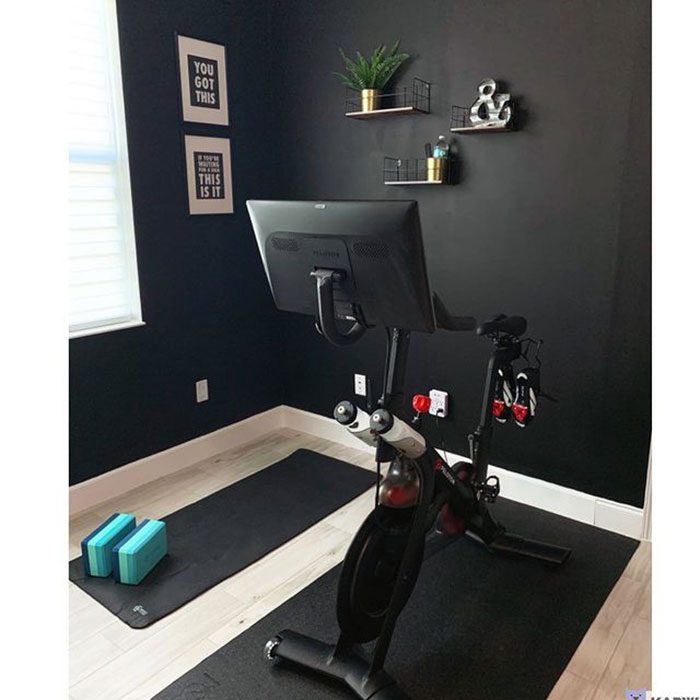 A stationary bike with a monitor stands on a mat in a stylish room with dark walls, a window, and decorative shelves.
