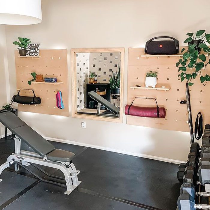 A workout bench sits in a well-lit room, surrounded by organized storage for fitness equipment, plants, and a mirror reflecting additional greenery.