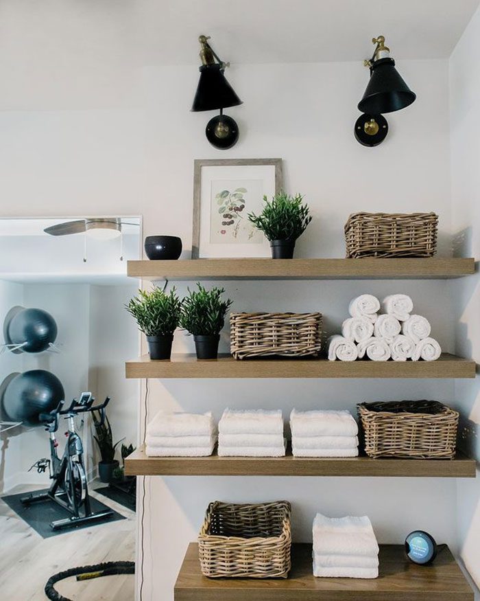 Shelves display neatly arranged towels and wicker baskets alongside potted plants and decor, with fitness equipment visible in the background of a well-lit room.