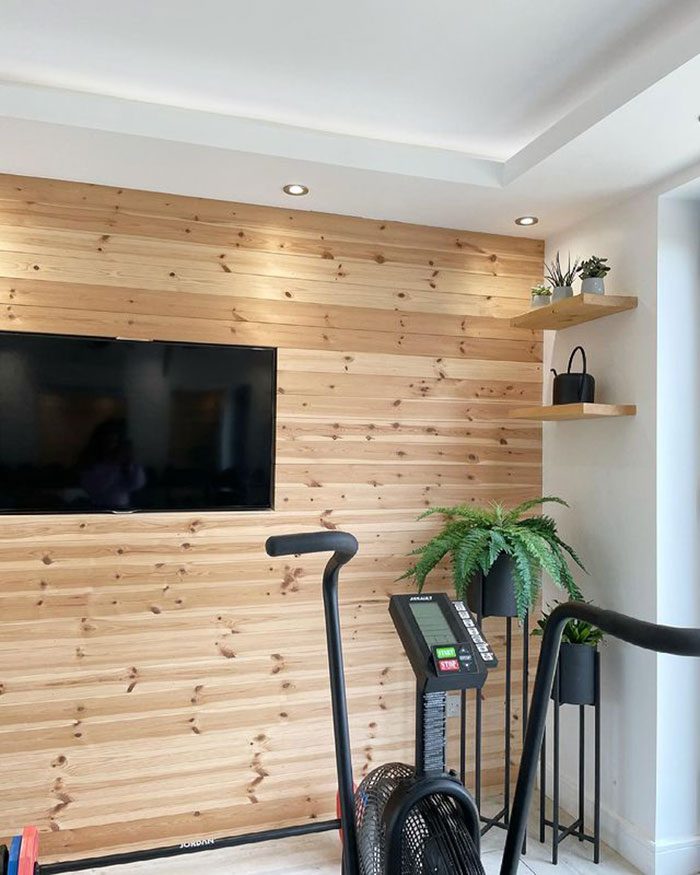 A wooden wall features a large television and shelves with plants. In front, exercise equipment is positioned, enhancing the modern, minimalist room's fitness atmosphere.