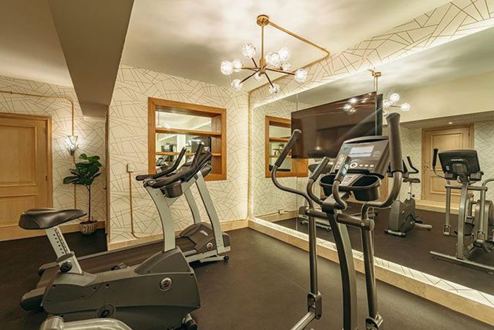 An exercise room contains an elliptical machine and stationary bike, illuminated by modern lighting, with a mirrored wall and decorative geometric wallpaper.