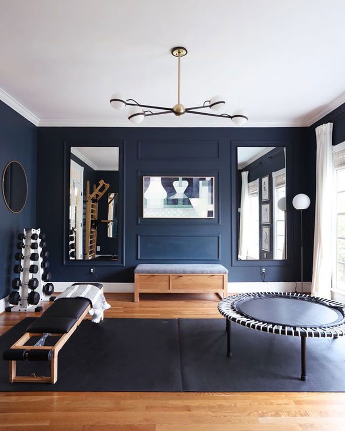A dark blue home gym features a bench, trampoline, and weights, illuminated by a modern chandelier, with mirrors reflecting the space's stylish design.