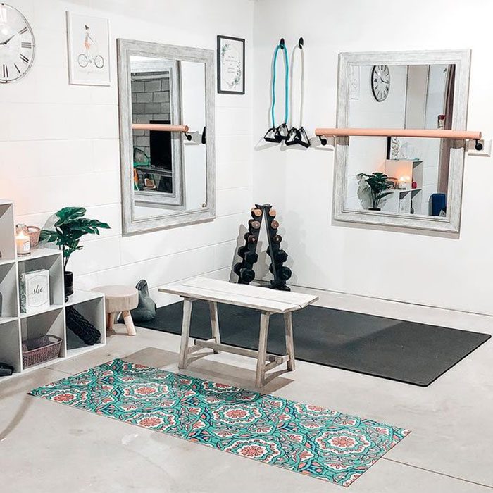 A wooden bench sits on a decorative mat in a minimalistic workout space with mirrors, weights, and fitness equipment against white walls.