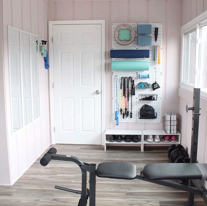 A workout bench occupies the center of a light-colored room, with fitness equipment neatly organized on a wall and shoes stored below.