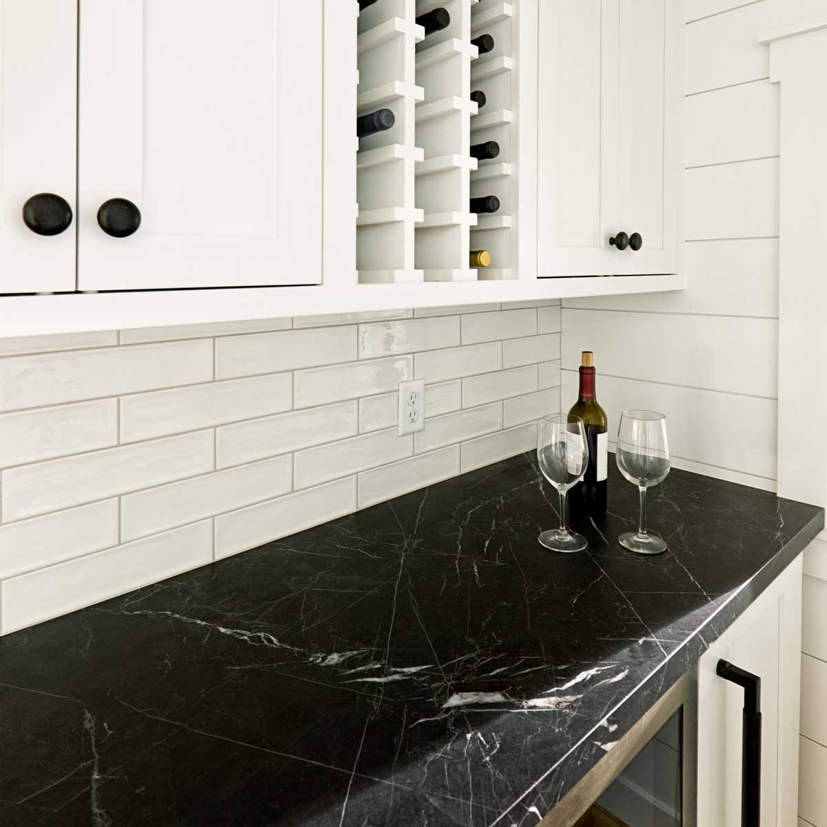 A black marble countertop holds a wine bottle and two glasses, situated below white cabinets and a tiled backsplash in a modern kitchen setting.