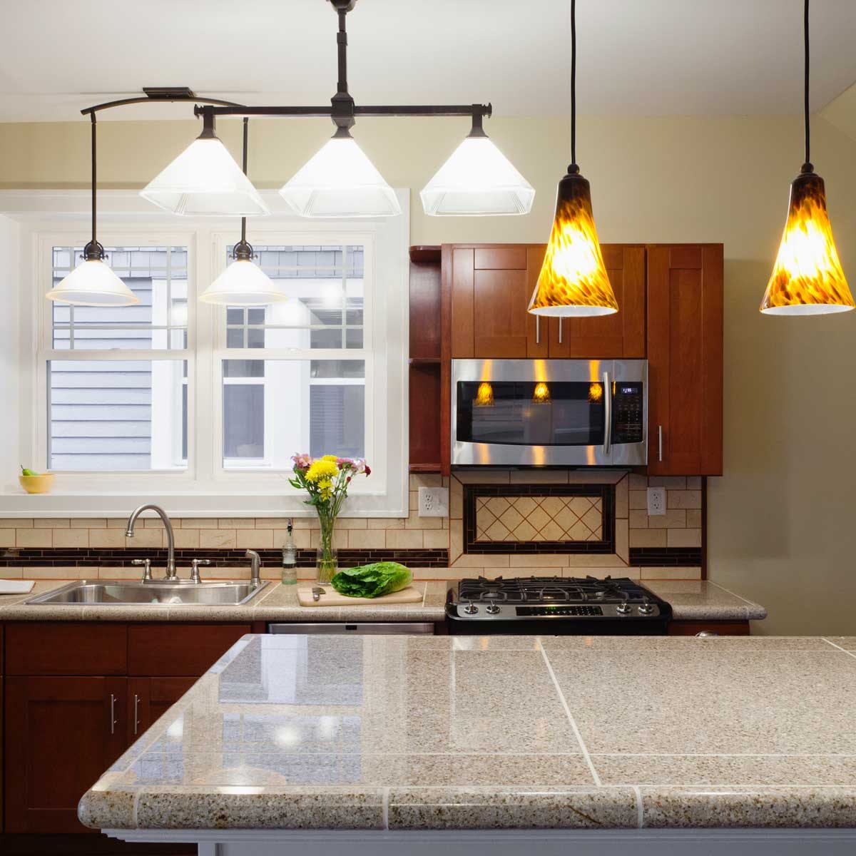 A modern kitchen features a countertop with fresh vegetables, cabinets, a microwave, and decorative pendant lights above, illuminated by natural light from the windows.