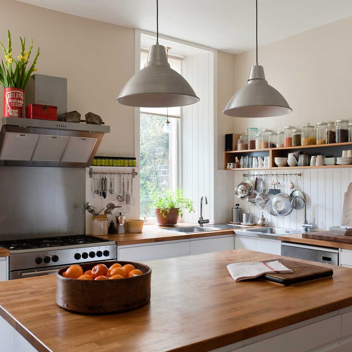 A wooden kitchen table holds a bowl of oranges. Above, two pendant lights hang. Shelves display jars and kitchenware in a bright, modern kitchen.