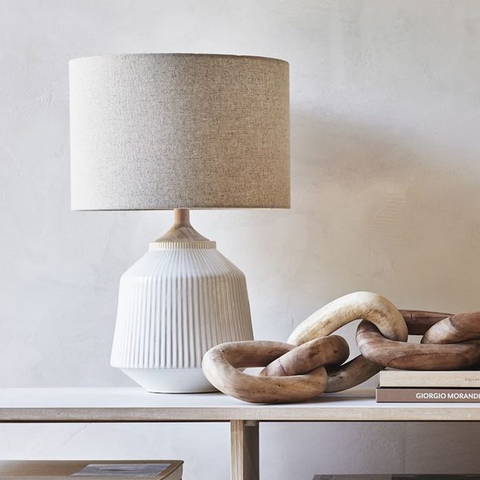 A decorative lamp stands on a wooden table, casting light, alongside intertwined wooden chains and a stacked book, against a neutral wall.