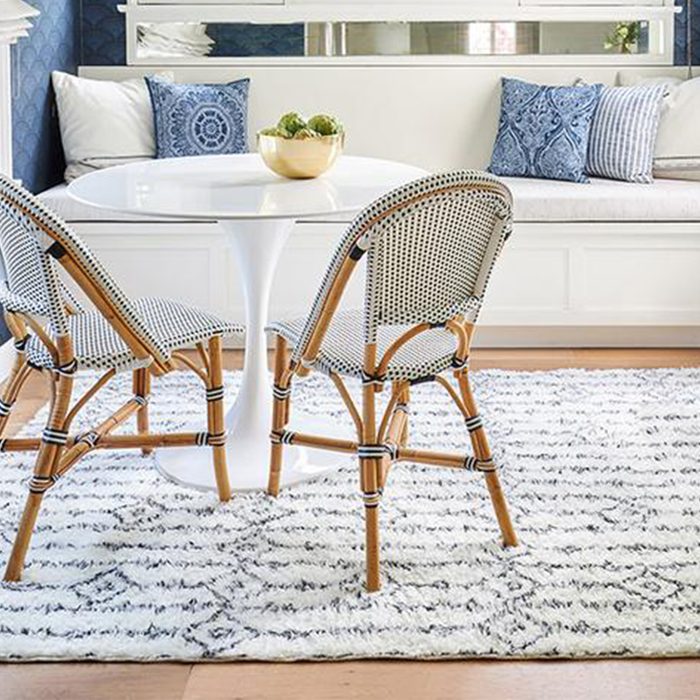 A round white table sits on a patterned rug, surrounded by two rattan chairs, with blue pillows and a bowl of decorative items in the background.