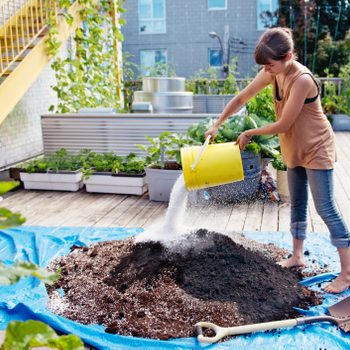 A woman pours soil from a yellow bucket onto a mound covered by a blue tarp, surrounded by a garden on a wooden deck.