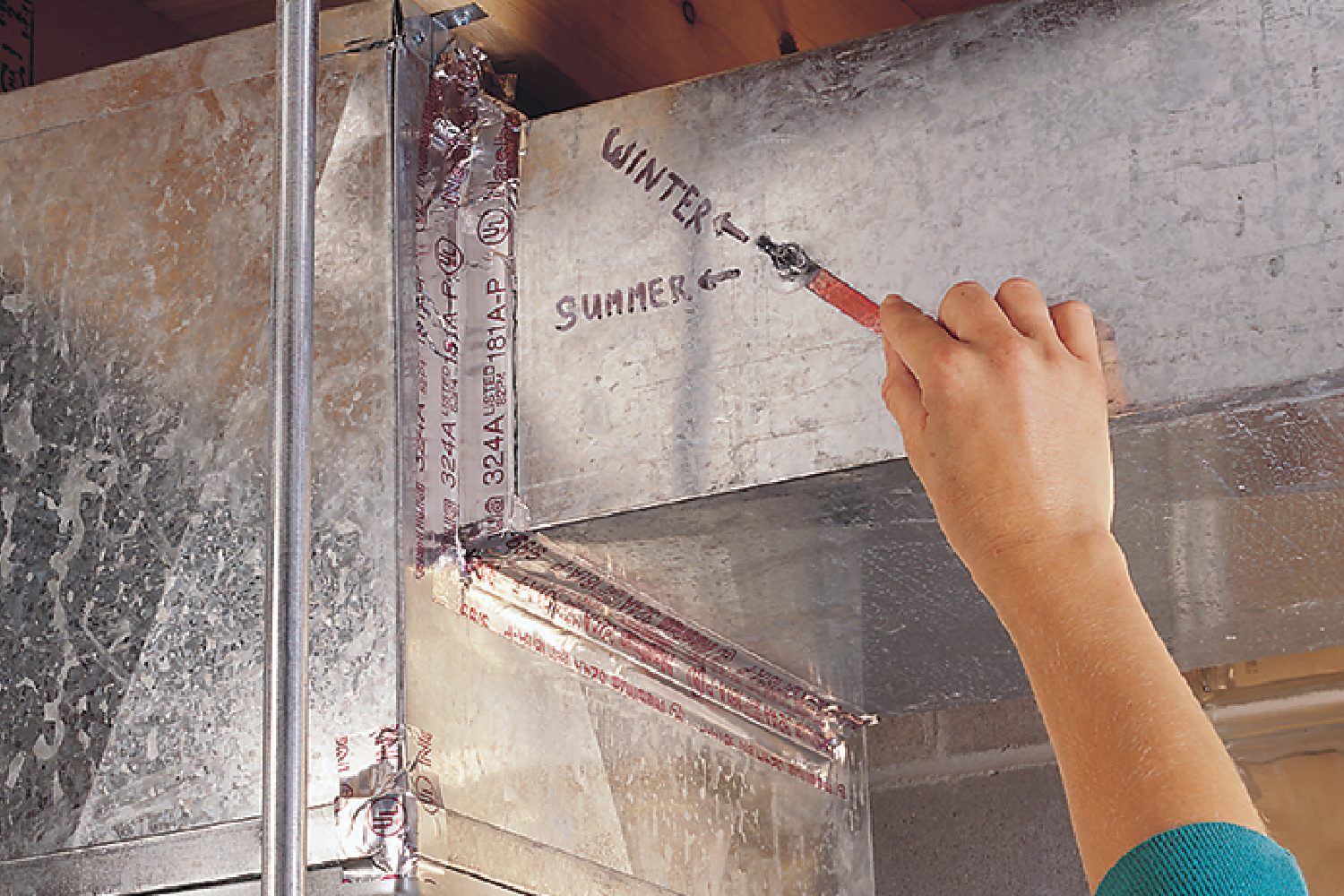 A hand uses a marker to label metal ductwork with "WINTER" and "SUMMER" while surrounded by insulation and structural elements in a building.