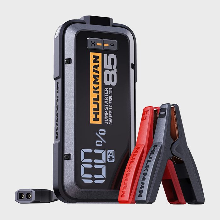 A portable jump starter with digital display shows battery percentage. Red and black clamps are attached, designed for automotive use.