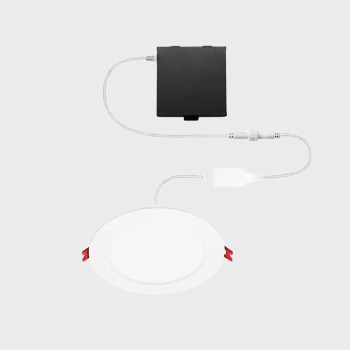 A round light fixture with red clips is connected via cables to a black box. The background is plain and light-colored.