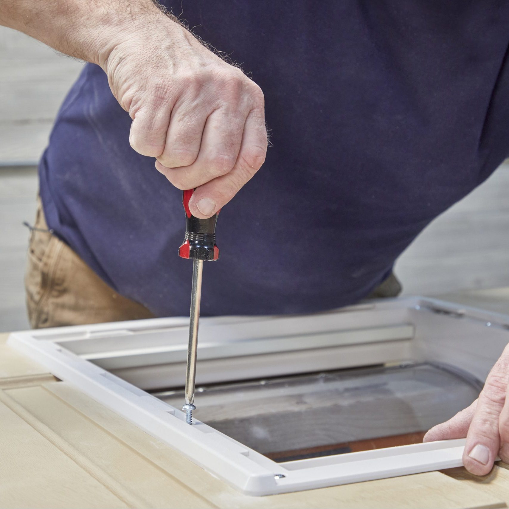 A hand turns a screwdriver, securing a screw into a white frame on a wooden surface, suggesting a repair or assembly task in a workshop.