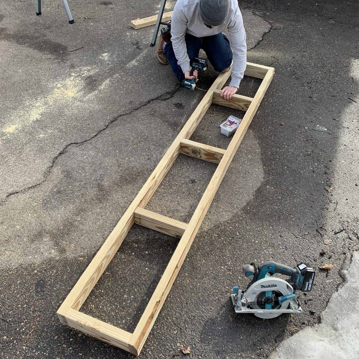 A person kneels on the ground, using a power drill to assemble a wooden frame on a paved surface, with a circular saw nearby.