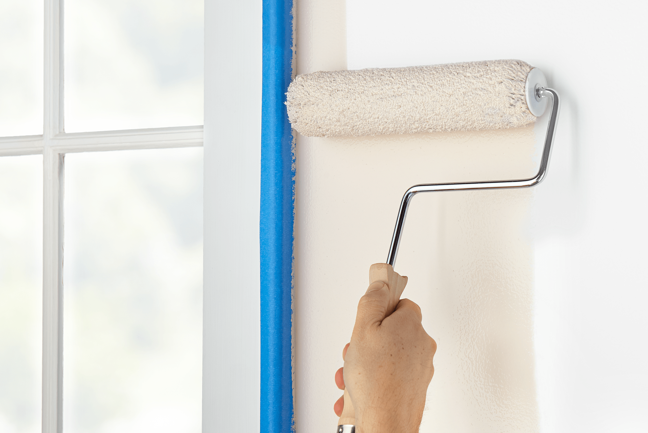 A paint roller held by a hand applies light-colored paint to a wall, framed by a blue tape, with a bright window in the background.