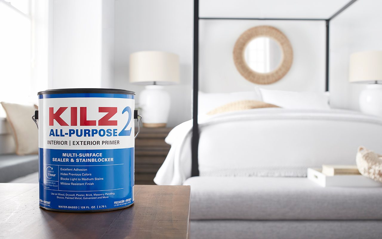 A blue paint can sits on a wooden table, showcasing a modern bedroom with a bright bed, lamps, and decorative circular mirror in the background.