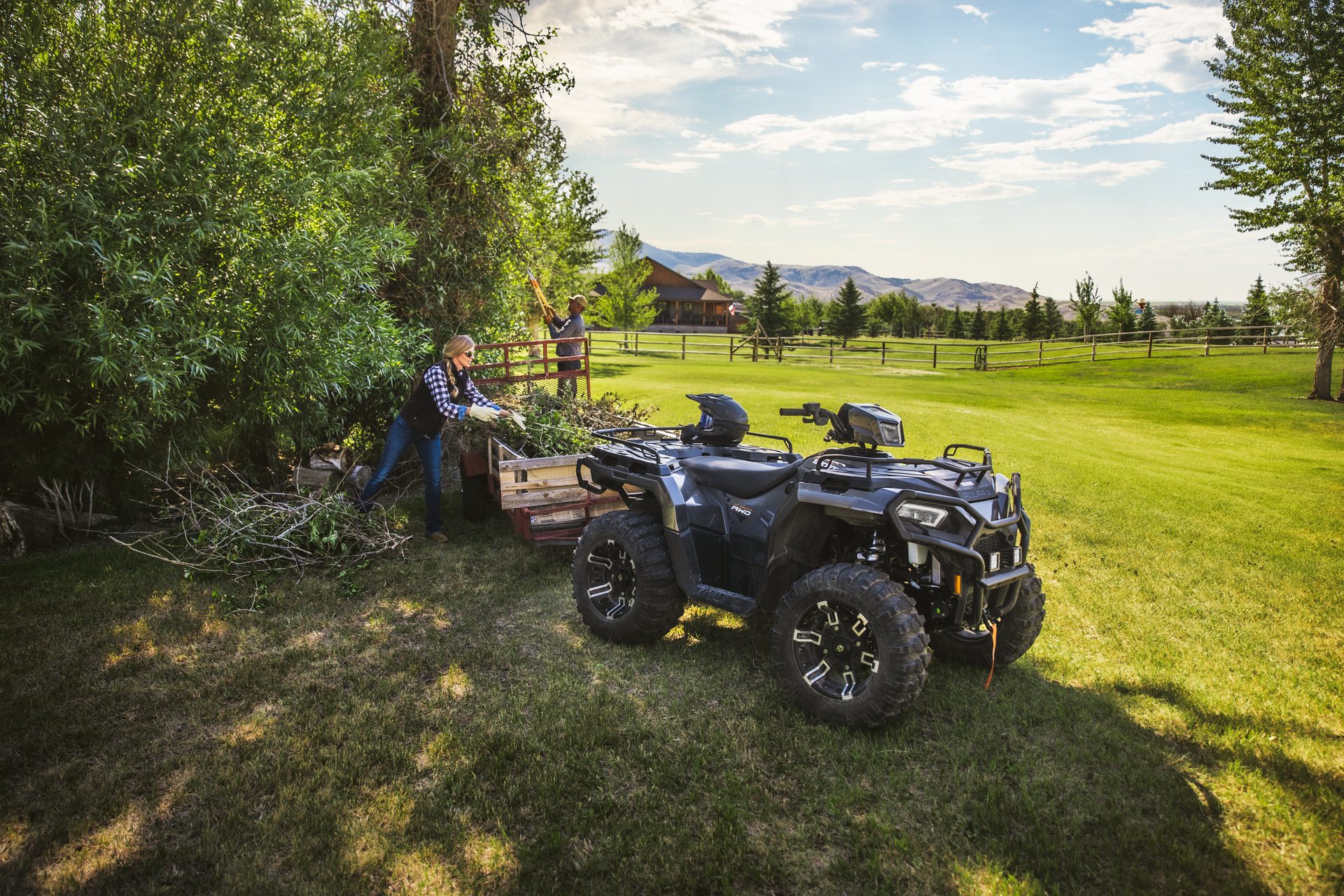 A woman pulls branches from a cart while a man trims trees nearby, with an ATV parked on grass in a scenic outdoor setting.