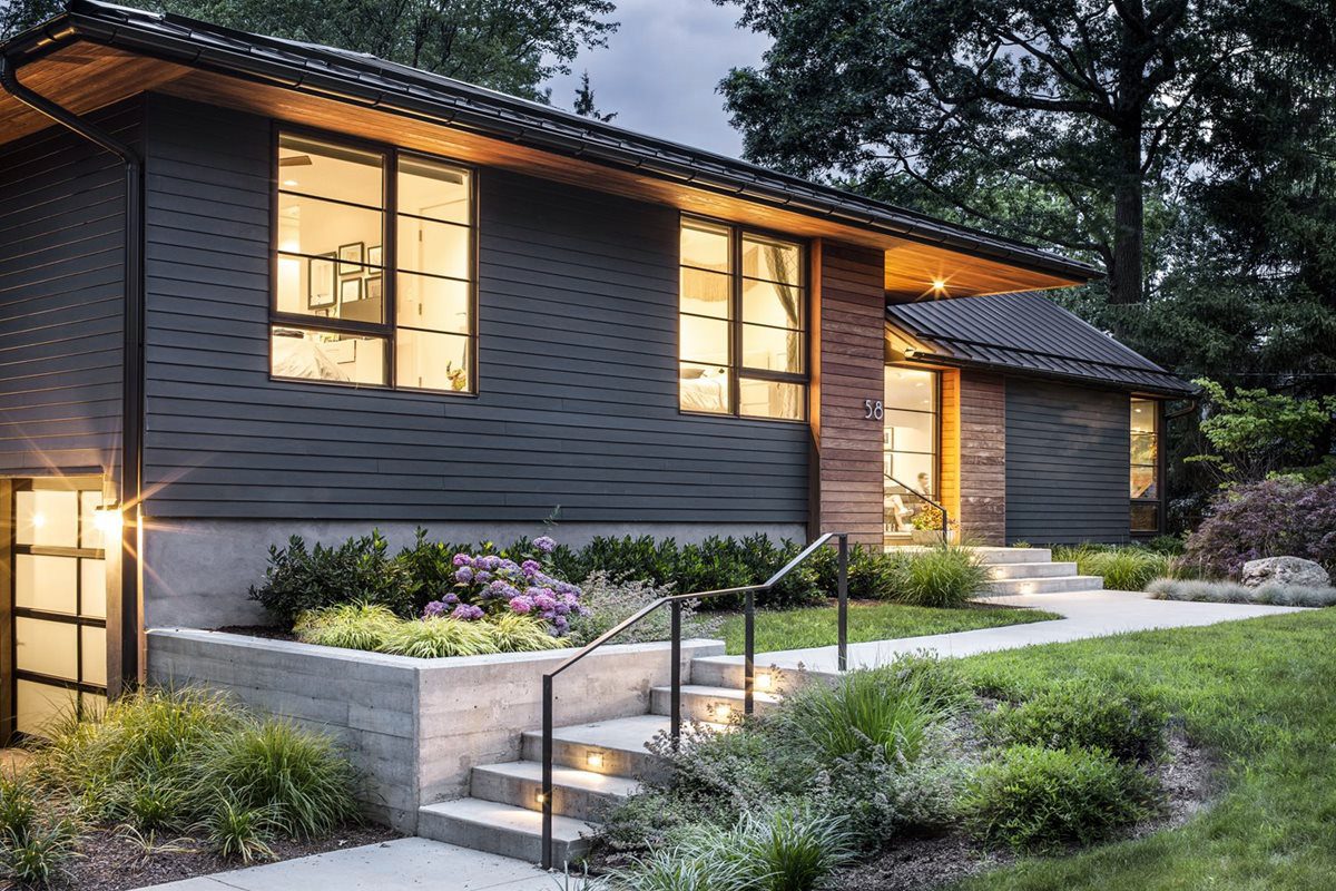 A modern house with large windows and porch lights stands amidst landscaped greenery and flower beds, illuminated softly during twilight.