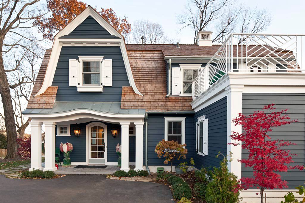 A stylish blue house with a steep roof showcases white shutters and a porch, surrounded by landscaping and autumn foliage under a clear sky.