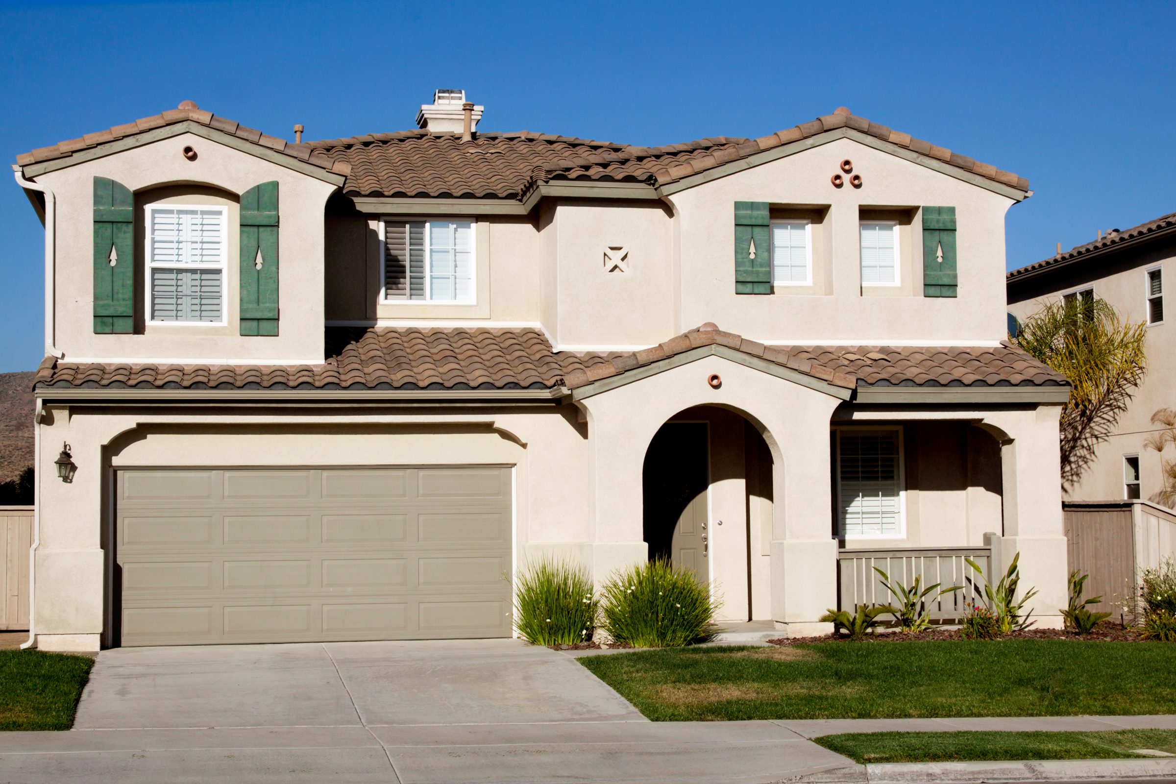 A two-story house features beige walls, green window shutters, and a tiled roof. It stands in a residential neighborhood under a clear blue sky.