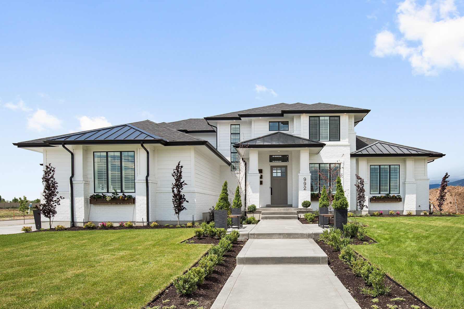 A modern white house with a black roof stands in a well-manicured yard, featuring a concrete pathway leading to a prominent entrance.
