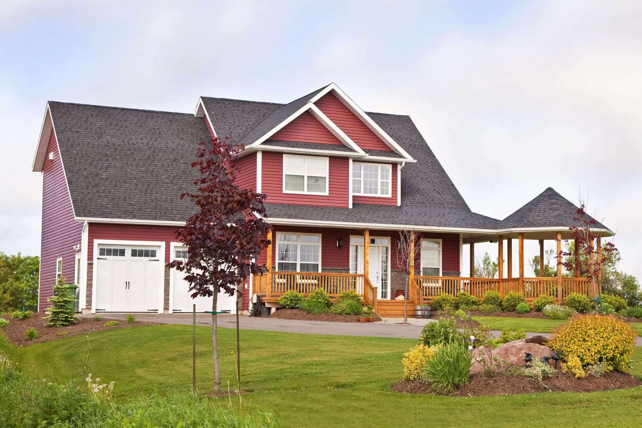 A red two-story house is situated on a grassy lawn, featuring a porch, trees, and flower beds, under a partly cloudy sky.