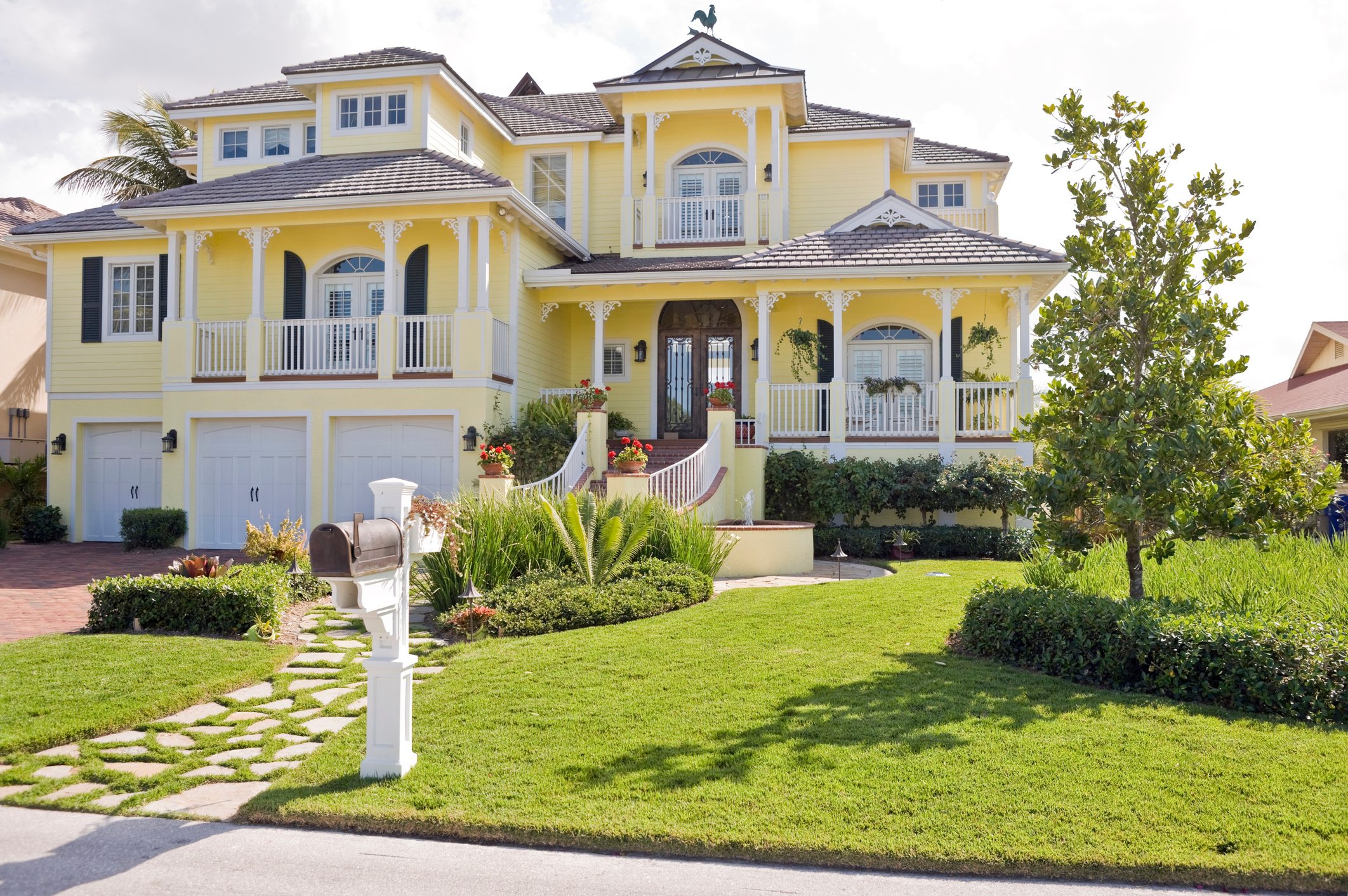 A yellow house with a decorative porch sits surrounded by landscaped gardens. A mailbox stands on a cobblestone path leading to the entrance.