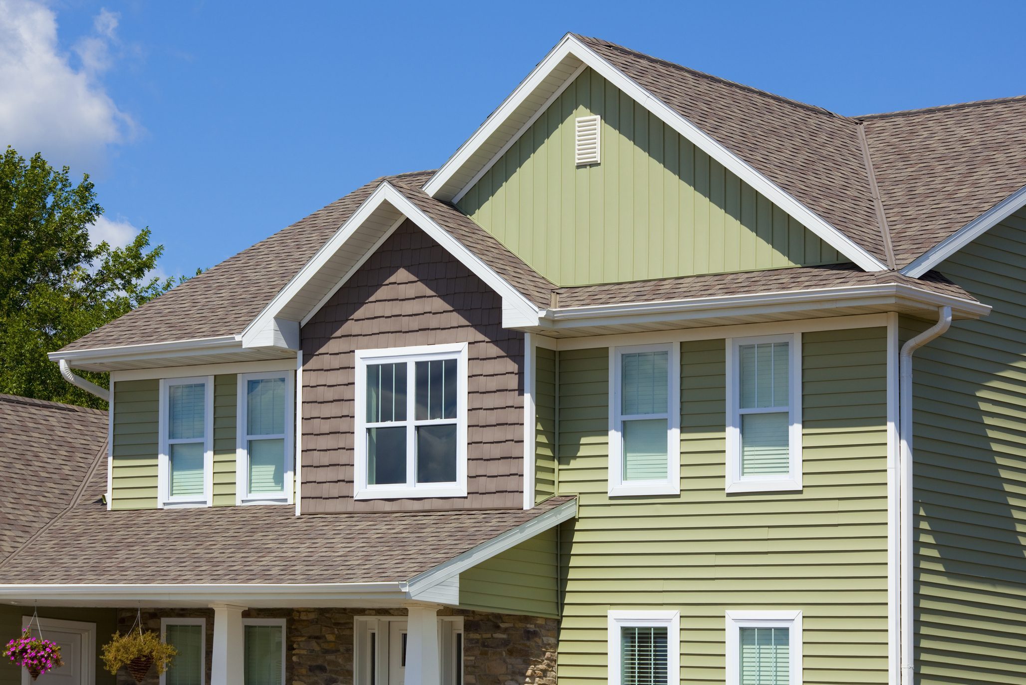 A multi-colored home stands under a clear blue sky, showcasing varied siding and roofing textures, with greenery and landscaping nearby.