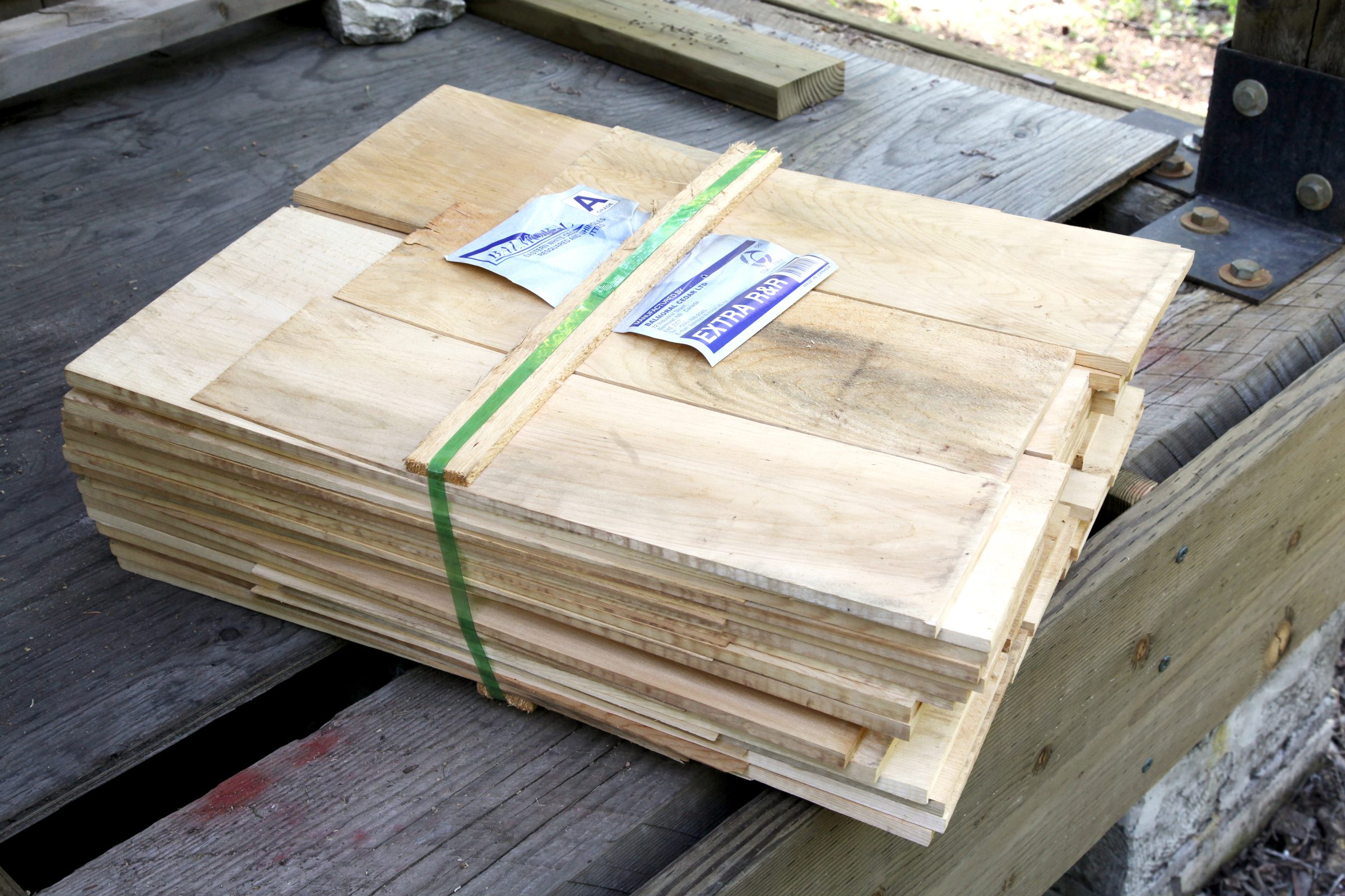 A stack of wooden boards is secured with green tape on a wooden surface, surrounded by other wood pieces and in a natural outdoor setting.