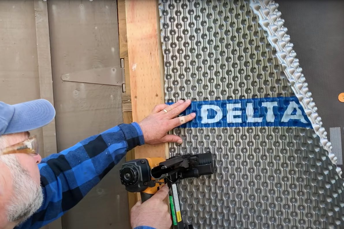 A hand holds a nail gun, fastening a textured panel labeled "DELTA" to wood in a construction setting.