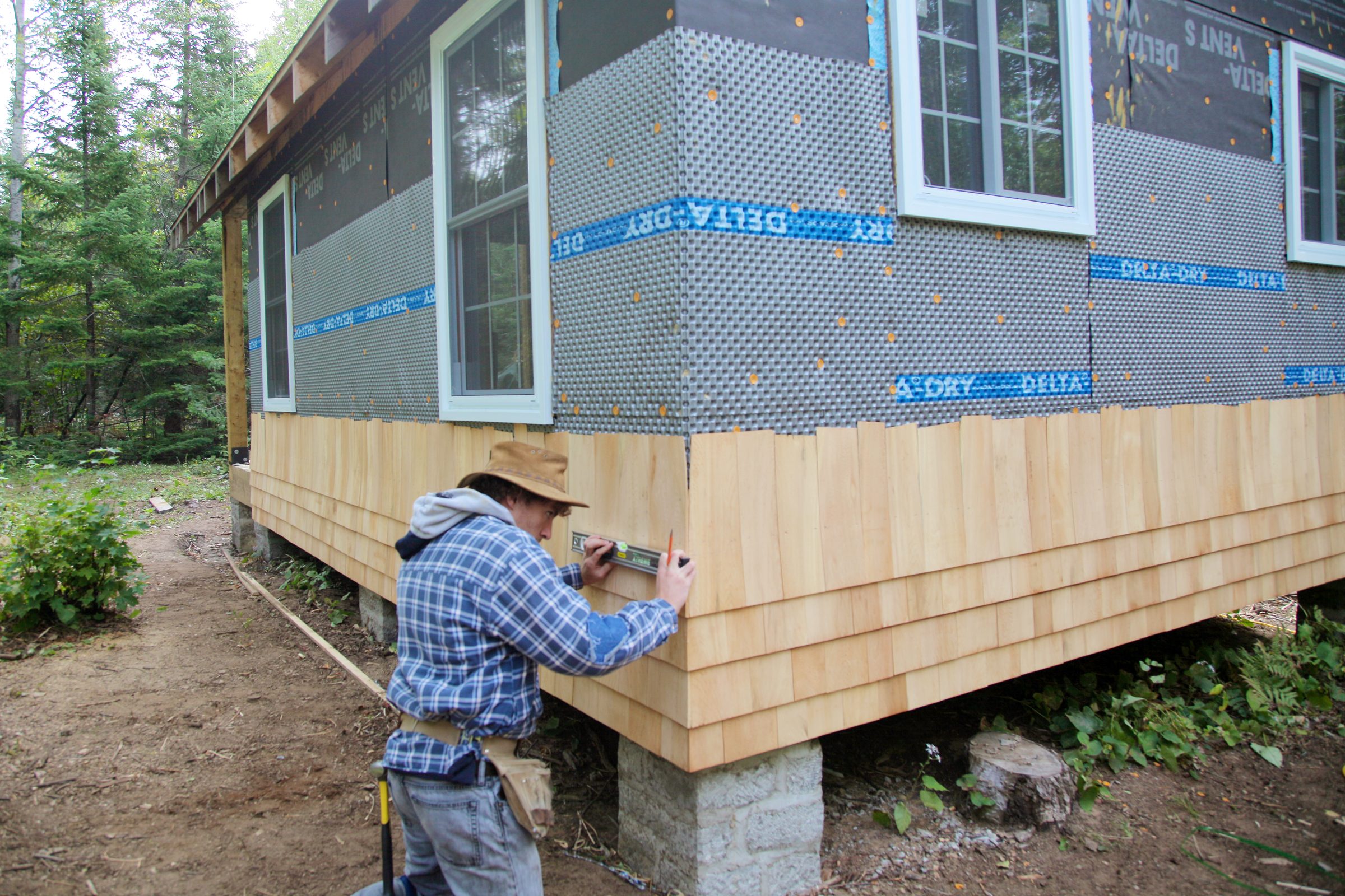 A person is measuring and installing wooden siding on a house in a wooded area, with a partially completed exterior visible in the background.